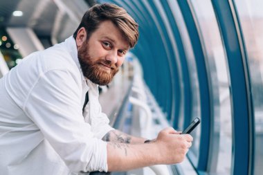 Modern cep telefonu cihazını halka açık bir yerde dinlenirken kameraya bakan, cep telefonuyla sohbet eden, cep telefonlu yakışıklı hippi portresi.