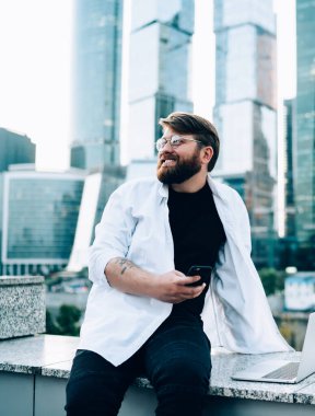 Gözlüklü, beyaz tişörtlü, cep telefonuyla mesajlaşan mutlu sakallı bir adam. Gökdelenleri olan bir barajın yanında duruyor.