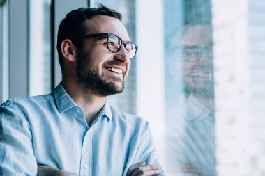Ofis penceresinin yanında elleri çapraz duran ve iş gününde gülümseyen neşeli erkek girişimci, Kafkasyalı başarılı şirket patronu zengin yaşam tarzından memnun.