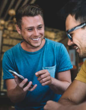 Mavi tişörtlü mutlu genç adam modern kafede buluşurken cep telefonuyla arkadaşına yeni bir uygulama gösteriyor.