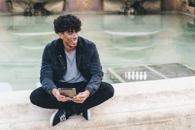 Genç, Afro-Amerikalı kıvırcık saçlı, fıskiyenin yanında bacak bacak üstüne atmış, elinde tabletle oturmuş başka tarafa bakan bir adam.