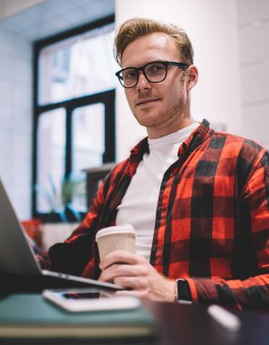 Aşağıdaki akıllı, sakin, odaklanmış erkek kahve kupası ile kafede mesai sırasında laptopta sörf yaparken kameraya bakıyor.
