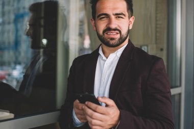 Sakallı, takım elbiseli İspanyol patron pencereye yaslanırken kameraya bakıyor ve modern binanın dışındaki akıllı telefona bakıyor.