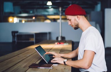 Odaklanmış profesyonel hippi yetişkin işçi ahşap masada oturmuş çatı katında dokunmatik ekranlı bir defter kullanıyor.