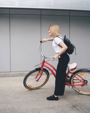 Modern binanın dış tarafına bakan pembe bisikletle gezerken kısa saçlı, şık sarışın bir kadının yan görünüşü.
