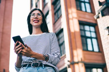 Yeni yayınlar yaratırken cep telefonlu ve gülümseyen mutlu milenyum blogcusu bakış açısının altında, olumlu bir kadın çevrimiçi sörf için cep telefonu kullanıyor.