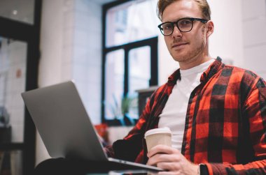 Aşağıdaki akıllı, sakin, odaklanmış erkek kahve kupası ile kafede mesai sırasında laptopta sörf yaparken kameraya bakıyor.