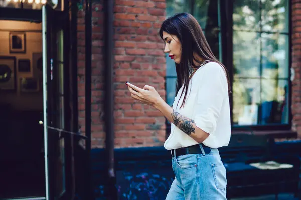 Side view of millennial female blogger installing medial application to smartphone device for editing photos using 4g wireless internet during walking time at city street, generation Y typing content