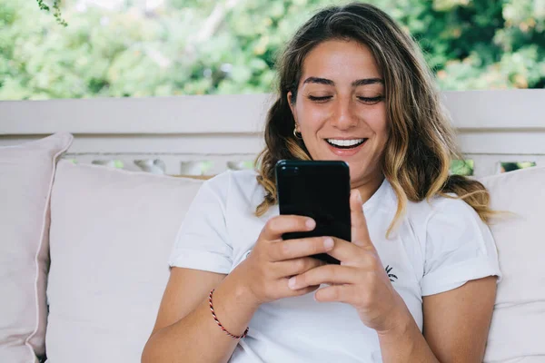 幸せな若い女性が携帯電話を使用して テラスで枕でソファに座っている間 ソーシャルメディアからのニュースに興奮笑っています — ストック写真