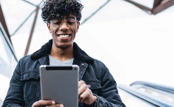 Optik gözlüklü neşeli Latin adam yürüyen merdivende aşağı iniyor ve dokunmatik ped aracılığıyla online sohbetten heyecanlanıyor, milenyum pozitif erkek blogcu mesajlar alıyor