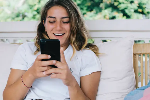 幸せな若い女性が携帯電話を使用して テラスで枕でソファに座っている間 ソーシャルメディアからのニュースに興奮笑っています — ストック写真