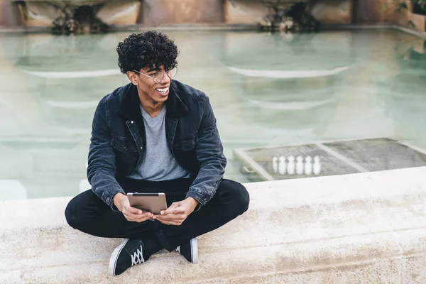 Genç, Afro-Amerikalı kıvırcık saçlı, fıskiyenin yanında bacak bacak üstüne atmış, elinde tabletle oturmuş başka tarafa bakan bir adam.