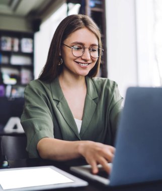 Kütüphanede dizüstü bilgisayarı ve tableti olan gözlüklü zeki ve neşeli öğrenci bulanık arka planda gülümseyip internette sörf yapıyor.
