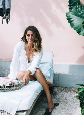 Plaj sandalyesinde oturmuş, kameraya bakarken dinlenen ve içtenlikle gülümseyen beyaz ceketli genç bir kadın.