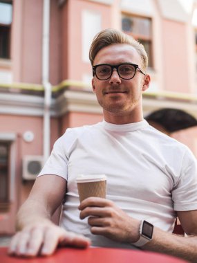 Tatmin olmuş beyaz gömlekli ve gözlüklü genç adam güneşli bir günde kafede dinlenirken bir fincan kahve içiyor.