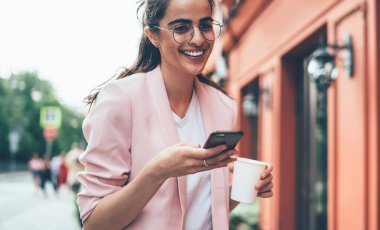 Düşük açılı, gözlüklü, sıcak içecekli zarif bir kadın cadde boyunca yürürken kameraya bakıp güler ve kırmızı rengin bulanık dış yüzüne karşı akıllı telefonla iletişim kurar.