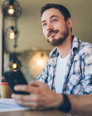 İş yerinde cep telefonuyla sörf yaparken kareli tişörtlü, güler yüzlü, düşünceli, düşünceli bir erkeğin bakışlarının altından.