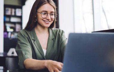 Kütüphanede dizüstü bilgisayarı ve tableti olan gözlüklü zeki ve neşeli öğrenci bulanık arka planda gülümseyip internette sörf yapıyor.