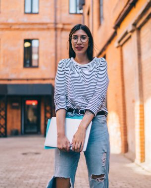 Serbest çalışmak için modern dizüstü bilgisayarı tutan ve boş zamanlarında açık havada kameraya bakan gündelik kıyafetli başarılı bir kadının yarım boy portresi.