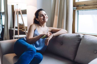 Kolsuz tişörtlü, kot pantolonlu, kanepede dinlenen, bilezik takan ve daireye bakan gülümseyen genç bayan.