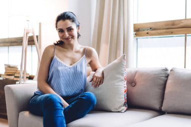 Kolsuz tişörtlü ve kot pantolonlu genç bir bayan bacak bacak bacak üstüne atmış, dirseğini yastığa yaslayıp gülümsüyor ve kameraya bakıyordu.