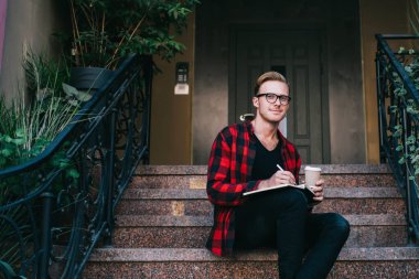 Yaratıcı gülümseyen kızıl saçlı adam kırmızı ve siyah gömlekli işaretli not defterine not alıyor ve yazın granit merdivenlerde kahveyle birlikte oturuyor.