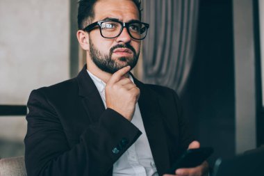 Resmi takım elbiseli ve gözlüklü düşünceli bir adam. Ortağıyla akıllı telefon mesajlaşması yapıyor. Çenesine dokunuyor.