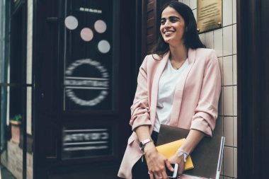 Beyaz gömlekli, pembe ceketli, akıllı telefon ve belgeli genç bir kadın girişte durup gülerken ve duvara yaslanırken.