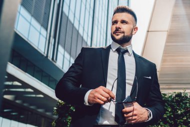 Düşük açılı, koyu renk takım elbiseli patron güneş gözlüğü ve akıllı telefon taşıyor ve şehir caddesindeki modern binaların dışındaki çalıların yanında dikilirken yüzünü çeviriyor.