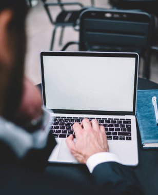 Yukarıdan gelen koyu renk takım elbiseli düşünceli yönetici, masada dizüstü bilgisayarda beyaz, tek kullanımlık defter ve kalemle yazı yazıyor.