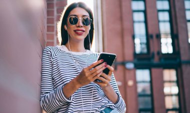 Modern cep telefonuyla radyo uygulaması, pozitif kadın kulaklıkları test eden modern cep telefonuyla şehir merkezinde arkadaşının müzik podcasının keyfini çıkaran bin yıllık hipster kızının yarım boy portresi.