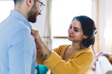 Gülümseyen sarı kazaklı genç bir kadın, işe gitmeden önce sevgili kocasını kolluyor ve tasmasını düzeltiyor.