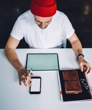 Yukarıdan gelen kırmızı şapkalı genç adam beyaz masada oturuyor ve tablet ve kırtasiye araçları ile cep telefonu kullanıyor.