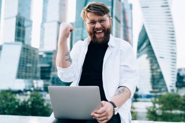 Saati ve dizüstü bilgisayarı olan, günlük kıyafetleriyle mutlu bir erkek iş bölgesinde çitlerin üzerinde başarılı bir projeyi kutluyor.