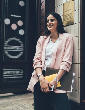 Beyaz gömlekli, pembe ceketli, akıllı telefon ve belgeli genç bir kadın girişte durup gülerken ve duvara yaslanırken. 