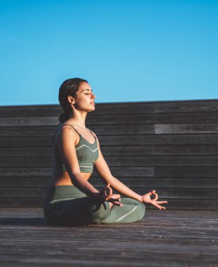 Yeşil spor elbiseli, sakin etnik zayıf bir kadının Lotus duruşu içinde el ele tutuşup ahşap terasta gözlerini kapamasının yan görüntüsü.