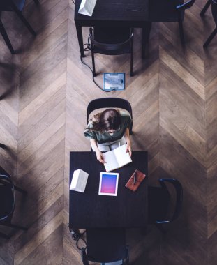 Yukarıdan gelen kadın, elinde kitap ve defterle dijital tabletin yanında oturmuş, gradyan ekranlı ve üniversite kütüphanesinde kitap okuyor.