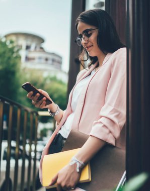 Kağıt klasörü ve günlüğü olan meşgul bir kadının cep telefonundan mesajları kontrol etmesi ve binanın yanında dururken gülümsemesi.