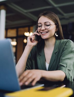 Güzel ve neşeli kız öğrenci kütüphanede dizüstü bilgisayarın önünde oturuyor ve sosyal medyada mesaj yazarken gülümsüyor ve akıllı telefondan konuşuyor.