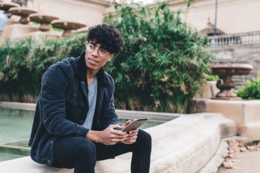 Gözlüklü ve elinde tabletle parkta çeşmenin kenarında oturan odaklanmış Afro-Amerikan bir adam.