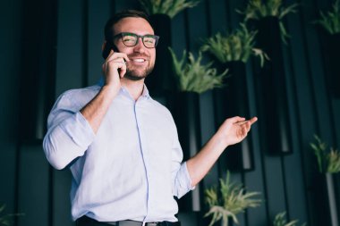 Modern ofisin koridorunda saksı bitkileriyle akıllı telefon konuşmaları yaparken gülümsemenin ve el kol hareketleri yapmanın düşük açısı.