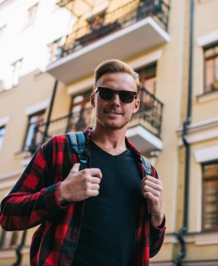 Güneş gözlüklü ve sırtında sırt çantası olan karizmatik yetişkin bir adam şehirdeki çok katlı binaların bulanık bej cephelerine gülümsüyor.