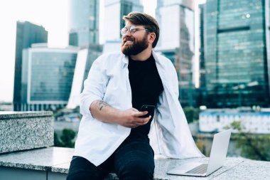 Gözlüklü, beyaz tişörtlü, cep telefonuyla mesajlaşan mutlu sakallı bir adam. Gökdelenleri olan bir barajın yanında duruyor.