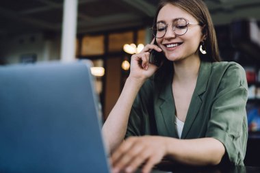 Güzel ve neşeli kız öğrenci kütüphanede dizüstü bilgisayarın önünde oturuyor ve sosyal medyada mesaj yazarken gülümsüyor ve akıllı telefondan konuşuyor.