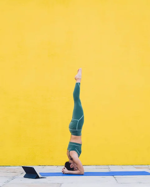 Side view of sportive slim woman in green outfit doing tripod headstand with legs raised straight up on yoga mat near tablet outdoors