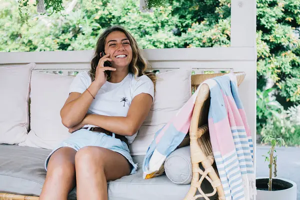 Joyful laughing young female talking on smartphone and sharing news with friend while sitting on wicker couch with pillows on terrace