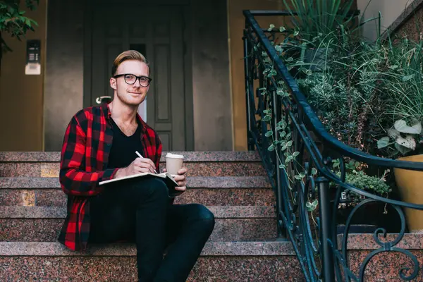 Yaratıcı gülümseyen kızıl saçlı adam kırmızı ve siyah gömlekli işaretli not defterine not alıyor ve yazın granit merdivenlerde kahveyle birlikte oturuyor.