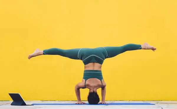 Back view of unrecognizable fit woman standing on mat in parivrtta eka pada sirsasana asana doing yoga with online class using tablet against yellow wall