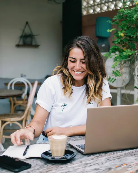 Cheerful young casual female reaching hand for pen for writing down information while sitting at old wooden table in cozy resort cafe and browsing internet on laptop