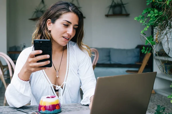 魅力的な女性で白いチュニックとともにDecolletサーフィンでインターネット上でラップトップと手に携帯電話を保持しながら 朝食を持っています — ストック写真